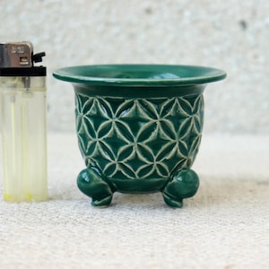 Può includere: Un piccolo vaso di ceramica verde scuro con un motivo geometrico e tre piedini. Il vaso ha un bordo largo ed è accanto a un accendino trasparente.