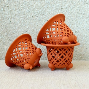 以下が含まれることがあります： テラコッタ色の透かし彫りセラミックプランター3つ。2つは横向きで、1つは直立しており、別のものと積み重ねられています。それぞれに装飾的な幾何学模様と小さな丸い足が付いています。