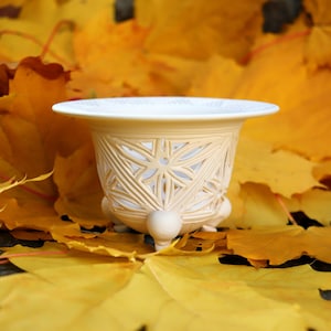 May include: A cream-colored, decorative planter with a geometric, openwork design. The planter sits on three small feet and has a wide rim. It is surrounded by vibrant yellow and orange autumn leaves, creating a seasonal display.