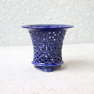 Può includere: Un vaso in ceramica blu cobalto con un motivo traforato. Il vaso ha una parte superiore svasata e una base arrotondata. L'intricato motivo traforato permette il passaggio della luce, creando un effetto decorativo. Il vaso è un elemento decorativo per la casa. Altezza circa 15 cm.