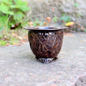 May include: A dark brown ceramic planter with a glossy finish and a textured, crisscross pattern on the exterior. The vessel features a wide, flared rim and is supported by three small, decorative feet at the base, creating a stable structure.