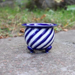 May include: A small, round ceramic planter featuring a glossy finish with a diagonal, spiraling pattern of deep blue and white stripes. The vessel has a wide, flared rim and is supported by three small, rounded blue feet at the base.
