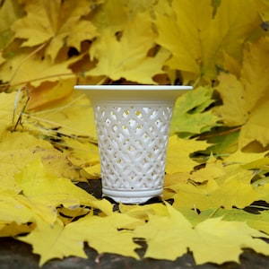 May include: A white, openwork ceramic vase with a geometric pattern. The vase is set against a backdrop of vibrant yellow autumn leaves. The vase has a wide rim and a detailed, lattice-like design.