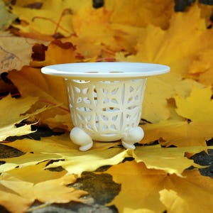 Può includere: Un vaso decorativo bianco in ceramica con un design traforato dettagliato. Il vaso ha un bordo largo e piatto e poggia su tre piccoli piedini arrotondati. È ambientato su uno sfondo di vivaci foglie autunnali gialle.