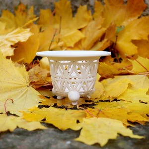 Peut inclure: Un petit panier décoratif blanc avec un motif en treillis, posé sur un lit de feuilles d'automne jaune vif. Le panier a un dessus rond et est soutenu par trois petits pieds arrondis.