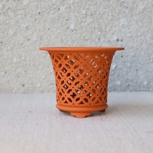 Op de afbeelding: Een oranje keramische plantenbak met een decoratief opengewerkt ontwerp. De plantenbak heeft een uitlopende bovenkant en een kleine, ronde basis met drie voetjes. Het ingewikkelde patroon zorgt voor luchtcirculatie en elegantie.