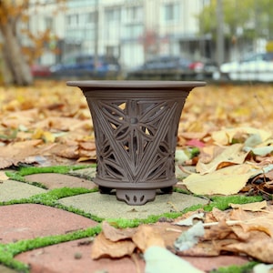 Può includere: Un vaso di ceramica marrone con un intricato design traforato. Il vaso ha una parte superiore ampia e una base decorativa. È appoggiato su una superficie di mattoni e pietra coperta di foglie cadute.