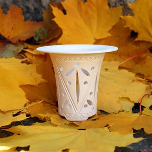 May include: A cream-coloured, decorative ceramic pot with a white rim, featuring cut-out designs. The pot is set against a backdrop of vibrant yellow autumn leaves, creating a seasonal aesthetic. The pot's height is approximately 13 cm.