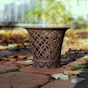 Può includere: Un vaso decorativo marrone con un design traforato, posto su una superficie di mattoni con foglie cadute. Il vaso ha una parte superiore svasata e una base dettagliata.