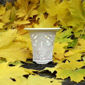 Puede incluir: Un jarrón de cerámica blanco, tallado intrincadamente con un diseño geométrico. El jarrón se encuentra sobre un fondo de vibrantes hojas de arce amarillas, creando un contraste de texturas y colores. El jarrón mide unos 10 cm de alto.