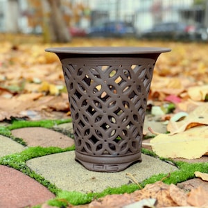 Può includere: Un vaso di fiori in ceramica marrone con un design traforato decorativo. Il vaso ha un bordo svasato e poggia su una base quadrata. Lo sfondo presenta foglie cadute e una superficie in mattoni e pietra.