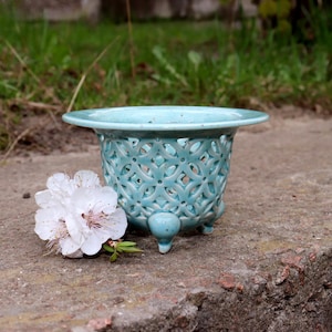 May include: A turquoise ceramic planter with a decorative openwork design and three small feet. A white flower with pink accents rests beside the planter. The planter is likely for small plants or succulents.