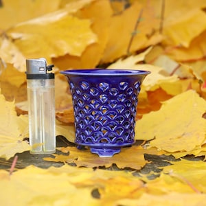 Può includere: Un vaso di ceramica blu cobalto con un design a griglia, accanto a un accendino trasparente. Il vaso è posto su un letto di foglie gialle autunnali, creando un contrasto di colori e trame.