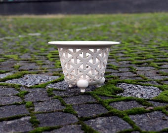 May include: A small, off-white ceramic bowl featuring an intricate, openwork lattice pattern across its exterior. The bowl has a wide, flat rim and is supported by three small, rounded ball-shaped feet at the base, creating a stable, elevated structure.