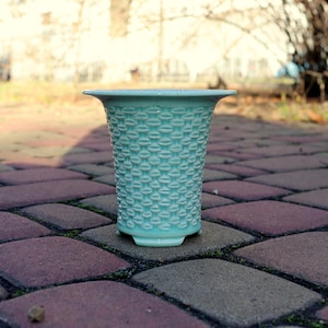 Può includere: Un vaso alto in ceramica verde acqua chiaro con un motivo strutturato e ripetuto. Il vaso ha un bordo svasato e una piccola base arrotondata. Il vaso è appoggiato su un patio in mattoni.
