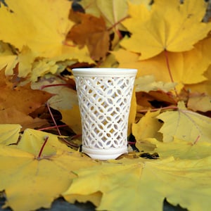 Könnte beinhalten: Eine cremefarbene Vase mit offenem Gitterwerk und geometrischem Muster. Die Vase steht vor einem Hintergrund aus leuchtend gelben und braunen Herbstblättern, was eine saisonale Darstellung ergibt. Das Design der Vase ist aufwendig.