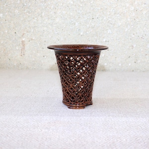May include: A dark brown ceramic vase featuring a wide, flared rim and a body constructed with an intricate, openwork lattice pattern of interlocking circles. The glazed finish provides a reflective surface across the entire exterior structure.