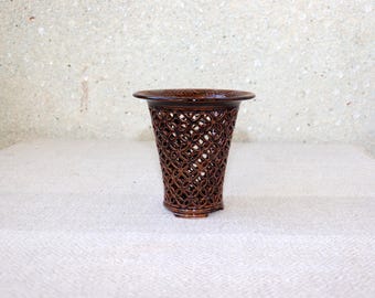 May include: A dark brown ceramic vase featuring a wide, flared rim and a body constructed with an intricate, openwork lattice pattern of interlocking circles. The glazed finish provides a reflective surface across the entire exterior structure.