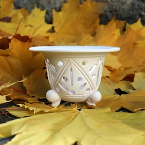 Op de afbeelding: Een crèmekleurige keramische plantenbak met een decoratief uitgesneden ontwerp, rustend op drie kleine voetjes. De plantenbak staat te midden van heldergele herfstbladeren, wat een seizoensgebonden scène creëert. De afmetingen zijn niet zichtbaar.