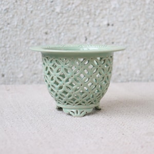 Puede incluir: Una maceta de cerámica verde pálido con un diseño calado decorativo. La maceta tiene un borde ancho y plano y se apoya en tres pequeños pies ornamentados. El patrón calado permite la circulación del aire, lo que la hace adecuada para plantas. La maceta mide aproximadamente 10 cm de alto.