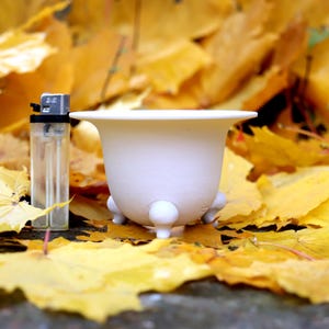 Può includere: Un vaso di ceramica bianco con tre piccoli piedini a sfera e un bordo svasato. Un accendino trasparente è accanto al vaso. Il vaso e l'accendino sono posti su uno sfondo di foglie gialle autunnali.