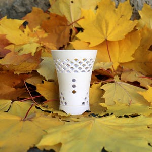 Può includere: Un vaso in ceramica bianca con un motivo decorativo traforato e fori circolari, su uno sfondo di vivaci foglie autunnali gialle. Il vaso ha una forma svasata e una piccola base.
