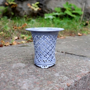 May include: A light periwinkle blue vase featuring an intricate, openwork lattice pattern throughout its cylindrical body. The vessel has a wide, flared rim at the top and a scalloped base, showcasing a detailed, repeating geometric design.