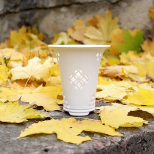 May include: A white ceramic candle holder with a flared rim and a tapered body featuring a central diamond-shaped cutout pattern and small circular perforations along the top and bottom edges, standing on a circular base.