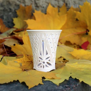 Può includere: Un portacandele in ceramica bianca finemente intagliato con un design geometrico e floreale. Il portacandele è posto su un letto di vivaci foglie autunnali gialle.