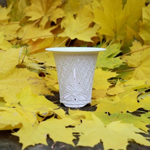 May include: A white, patterned ceramic vase with a detailed geometric design. The vase is set against a bed of bright yellow maple leaves, creating a contrast of textures and colours. The vase has a wide rim and a slightly flared base.