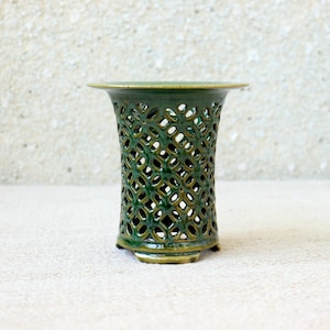 Peut inclure: Un vase en céramique vert foncé présentant un motif en treillis ajouré complexe sur tout son corps cylindrique. Le récipient possède un bord large et évasé au sommet et repose sur une petite base circulaire, avec une finition émaillée brillante.
