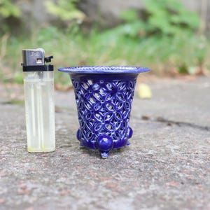 May include: A small, deep blue ceramic vessel with an intricate, openwork lattice pattern and a flared rim. The vessel stands on three small, rounded feet and is positioned next to a standard-sized rectangular lighter for scale comparison.