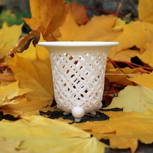 Può includere: Un vaso in ceramica traforata color crema, con bordo svasato e tre piccoli piedini a sfera. Il vaso presenta un motivo geometrico e si trova tra vivaci foglie autunnali gialle.