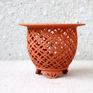 Può includere: Un vaso in ceramica color terracotta con un design traforato decorativo. Il vaso ha un bordo largo e poggia su tre piccoli piedini. Il motivo traforato è un disegno geometrico ripetitivo. Altezza di circa 15 cm.