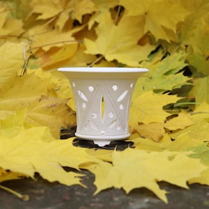 Pode incluir: Um castiçal de cerâmica branca com padrões recortados decorativos, sobre um fundo de vibrantes folhas de outono amarelas. O castiçal tem um topo alargado e uma base detalhada, ideal para decoração de casa.