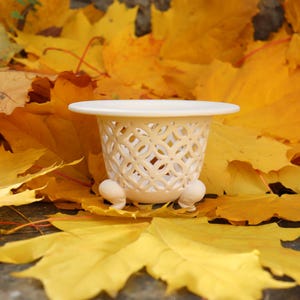 Può includere: Un piccolo vaso di ceramica bianco sporco con un design traforato decorativo e tre piedini. Il vaso è posto tra un letto di vivaci foglie autunnali gialle e arancioni, creando un'estetica calda e stagionale.