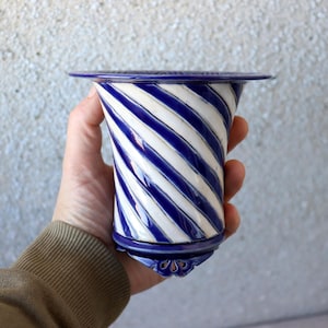 Può includere: Un vaso in ceramica blu e bianco con un design a spirale. Il vaso ha una parte superiore svasata e una base decorativa. Le strisce blu sono uniformemente distanziate e creano un motivo visivamente accattivante. Il vaso è tenuto nella mano di una persona.
