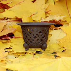 Può includere: Un vaso di ceramica marrone con un design traforato decorativo, appoggiato su un letto di vivaci foglie gialle autunnali. Il vaso ha un bordo svasato e tre piccoli piedini arrotondati. Le foglie creano un caldo sfondo stagionale.