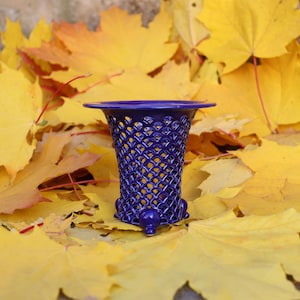 Può includere: Un vaso blu cobalto con un motivo a griglia, bordo svasato e piedini. Il vaso è posto su uno sfondo di vibranti foglie di acero gialle, creando un contrasto sorprendente di colori e trame. Arredamento per la casa.