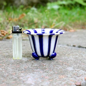 May include: A small ceramic planter featuring a white and dark blue vertical striped pattern with a flared rim and three rounded feet, positioned next to a standard disposable lighter for scale to show its compact size.