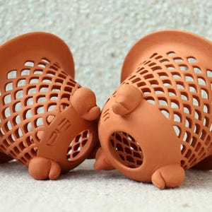 Puede incluir: Dos cestas de cerámica de color terracota. Cada cesta tiene un borde acampanado y un cuerpo con un patrón de celosía. Las cestas tienen pequeños pies redondeados y un elemento decorativo en el lateral.