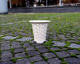 May include: A white ceramic vessel featuring a repeating geometric Y-shaped lattice pattern throughout its exterior. The object has a wide, flared circular rim at the top and a narrow base, showcasing a hollow, open-work design structure.