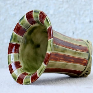 Può includere: Un vaso in ceramica fatto a mano con la parte superiore svasata e un corpo cilindrico. Il vaso presenta fasce alternate di smalto verde e marrone rossiccio. L'interno è di colore verde tenue. Il vaso è appoggiato su un lato.