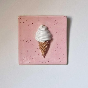 Op de afbeelding: Een vierkante, roze keramische tegel met een gespikkeld patroon. Een 3D-ijshoorntje is gecentreerd op de tegel. Het ijs heeft een witte werveling en de hoorn is goudbruin van kleur. De tegel is een decoratief item.