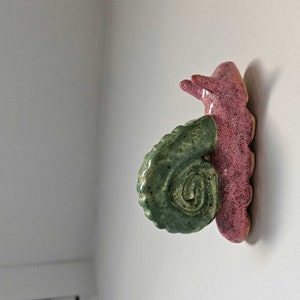 Op de afbeelding: Een keramische wandsculptuur met een gestileerd slakkenontwerp. De sculptuur heeft een roze, getextureerd lichaam en een groene, spiraalvormige schelp. Het kunstwerk is aan een witte muur bevestigd.