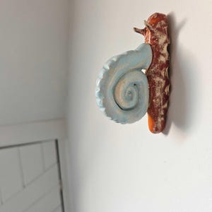 Può includere: Decorazione da parete in ceramica a forma di lumaca. Il guscio della lumaca è azzurro chiaro con un motivo a spirale. La lumaca è attaccata a una base marrone strutturata. La decorazione da parete è montata su una parete bianca.