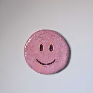 Könnte beinhalten: Eine rosafarbene Keramik-Smiley-Gesichtswanddekoration mit lila Glasur. Das Smiley-Gesicht hat ein einfaches Design mit zwei Augen und einem geschwungenen Mund.