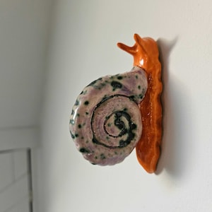 Op de afbeelding: Een keramische slaksculptuur met een spiraalvormig huis in tinten van lavendel, groen en wit. Het lichaam van de slak is oranje. De sculptuur is bevestigd aan een witte muur.