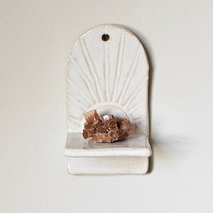Op de afbeelding: Een wit keramisch wandrek met een zonnestraalontwerp en een klein plankje. Een cluster van bruine kristallen rust op het plankje. Het rek heeft een gat aan de bovenkant om op te hangen. De algehele esthetiek is minimalistisch en natuurlijk.