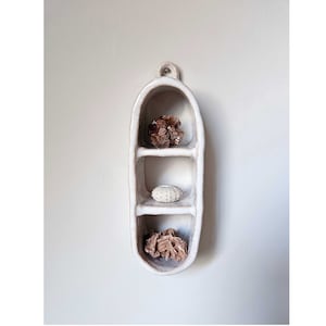 Op de afbeelding: Witte keramische wandplank met drie planken, gevormd als een peul, met een kleine bruine steen, een witte steen en een bruine steen op de planken.